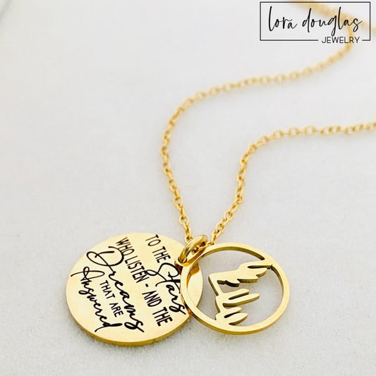 A gold stainless steel necklace with a round pendant engraved with the quote 'To the Stars who listen - and the Dreams that are Answered' and a laser cut mountain pendant.