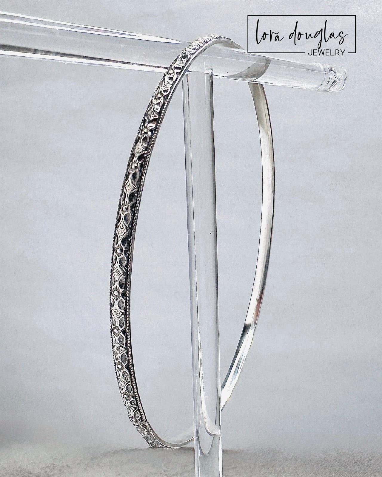 Handcrafted sterling silver bangle bracelet with textured and polished finish displayed on clear stand.