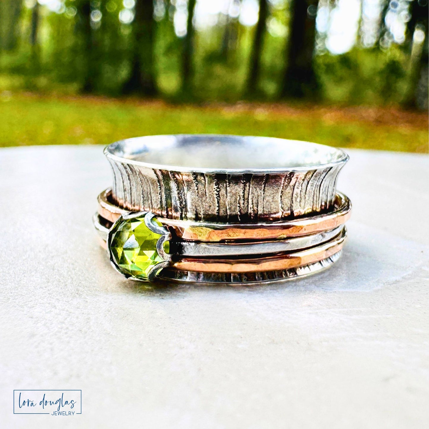 Stack of silver and rose gold rings with a green gemstone on a white surface with a blurred natural background.