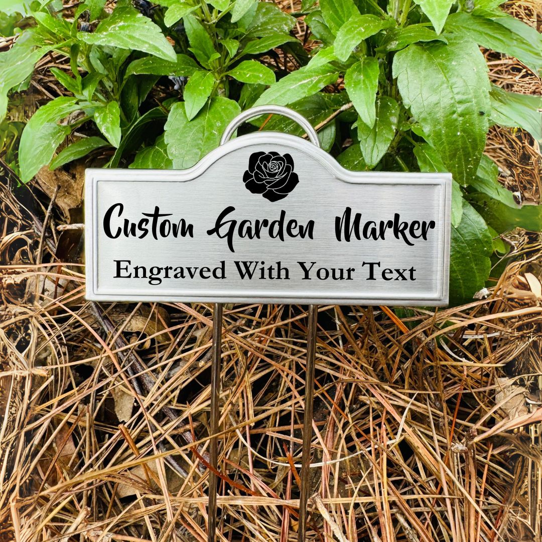 A stainless steel custom garden marker with engraved text and a floral design, placed in a garden setting.