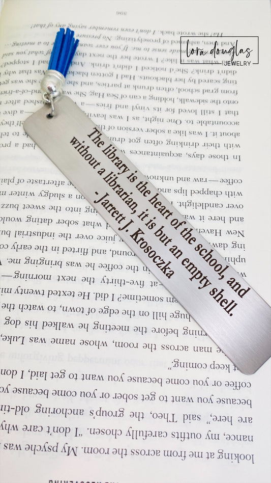 A stainless steel tassel bookmark with an engraved quote 'The library is the heart of the school...' placed on an open book.