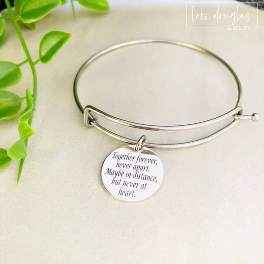A stainless steel adjustable bangle bracelet with a round pendant that has engraved text 'Together Forever, Never Apart, Maybe in Distance, But Never at Heart'.