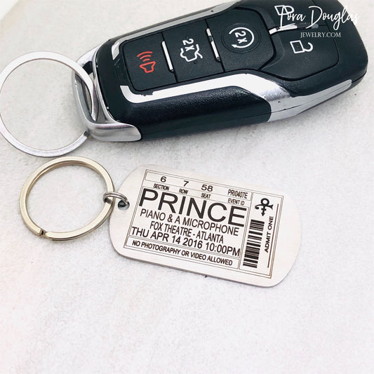 A stainless steel key chain with an engraved metal tag featuring text that resembles a Prince concert ticket. The key chain is shown with a car key.