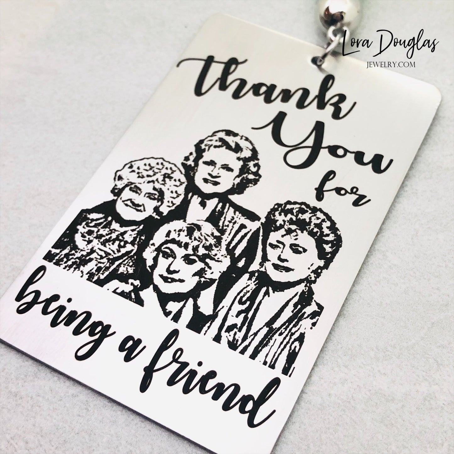 A stainless steel bookmark with engraved text and illustrations of the Golden Girls characters, with a navy blue velvet drawstring pouch and a gift tag.