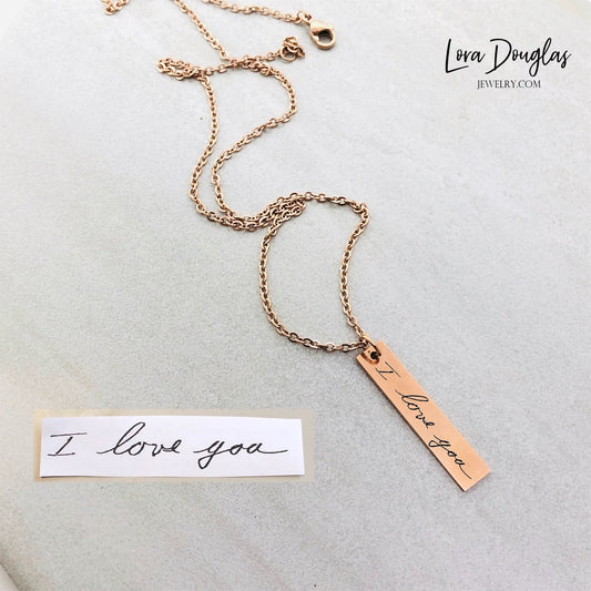 A gold stainless steel pendant with engraved handwriting, attached to an 18 inch chain with a lobster clasp, displayed against a white background with a blurred background hinting at a floral arrangement.