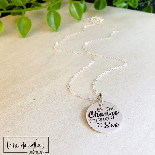 be the change you want to see necklace with 3/4 inch round engraved charm in stainless steel gold or silver finish displayed on chain highlighting minimal design and inspirational message jewelry