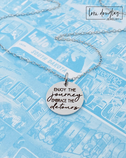 A silver necklace with a lobster clasp and a stainless steel bangle bracelet, both featuring an engraved message 'Enjoy the Journey, Embrace the Detours'