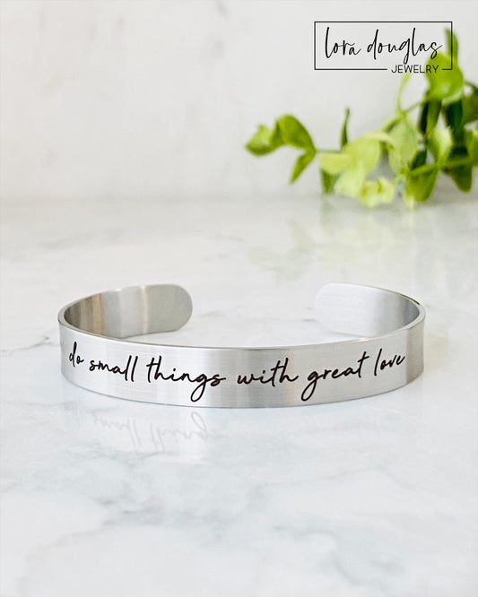 Two stainless steel cuff bracelets, one in silver and one in gold color, each with an engraved message 'Do small things with great love'.