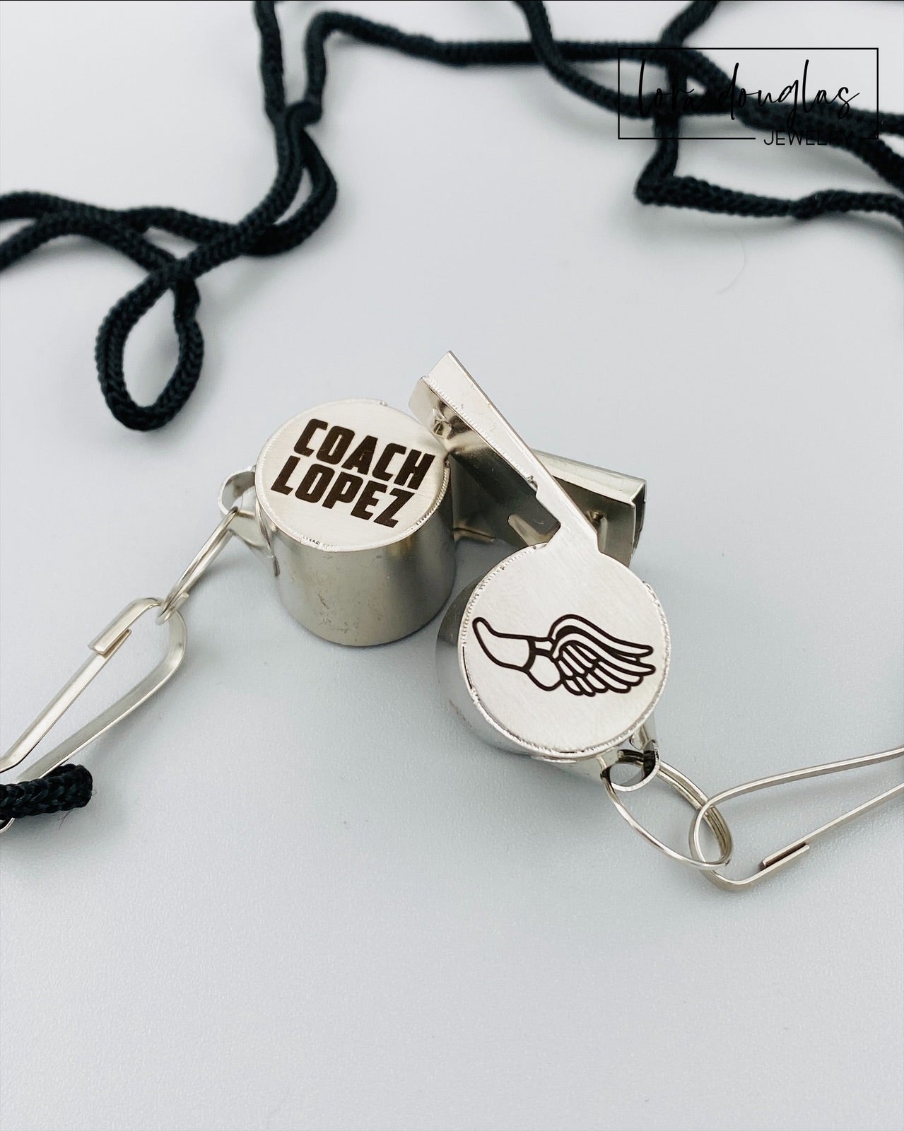 personalized stainless steel whistles with engraved text, one with 'COACH CARLOS' and sports icons, the other with 'FOOTBALL BASKETBALL SOCCER LIFEGUARD AND MORE'. Both whistles have a lanyard