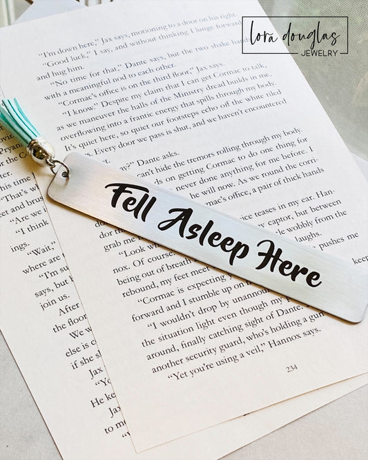 A stainless steel bookmark with the text 'Fell Asleep Here' engraved on it, displayed on an open book. The bookmark has a tassel and is attached to a navy blue velvet drawstring pouch with a gift tag.