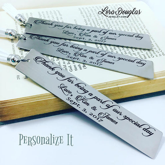 Two stainless steel bookmarks with personalized engraving, 'Thank you for being a part of our special day' and names, the other with a date. They are displayed on a white background with decorative tassels and flowers in the background.
