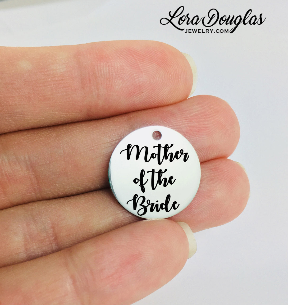 Round stainless steel pendant with the engraved text 'Mother of the Bride' held between fingers.