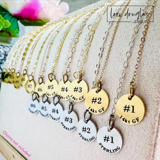 A display of Lora Douglas Jewelry Permanent Jewelry ID Tags for Displays, gold and silver metal labels numbered #1–#10 (“14kt GF” or “STERLING”), hangs on chains over a beige stand. Chains not included; flowers and branding in background.
