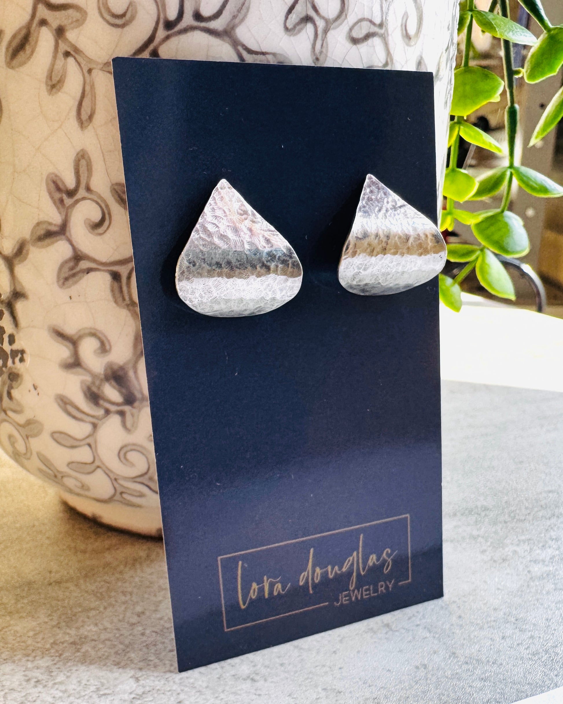 Teardrop-shaped sterling silver earrings on a Lora Douglas Jewelry card with a plant in the background.