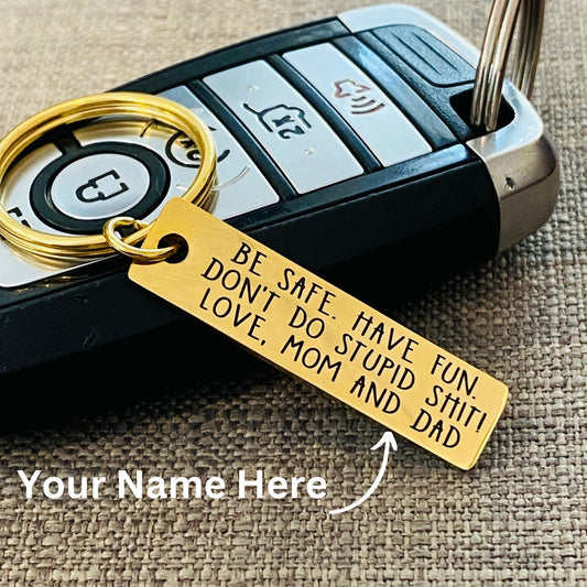 A gold metal keychain with a mirror finish, laser engraved with the message, Be Safe Have Fun Don't Do Stupid Shit. The keychain is displayed alongside a car key, and there is a placeholder for a name engraving.