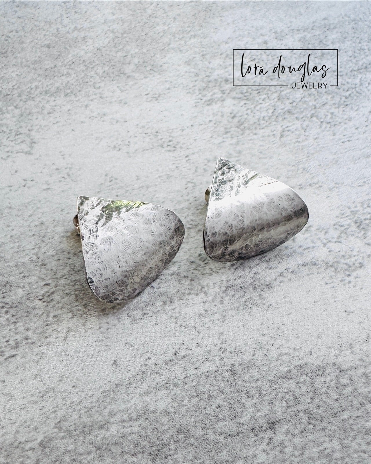 Pair of sterling silver earrings on a textured gray surface with 'Lora Douglas Jewelry' branding.
