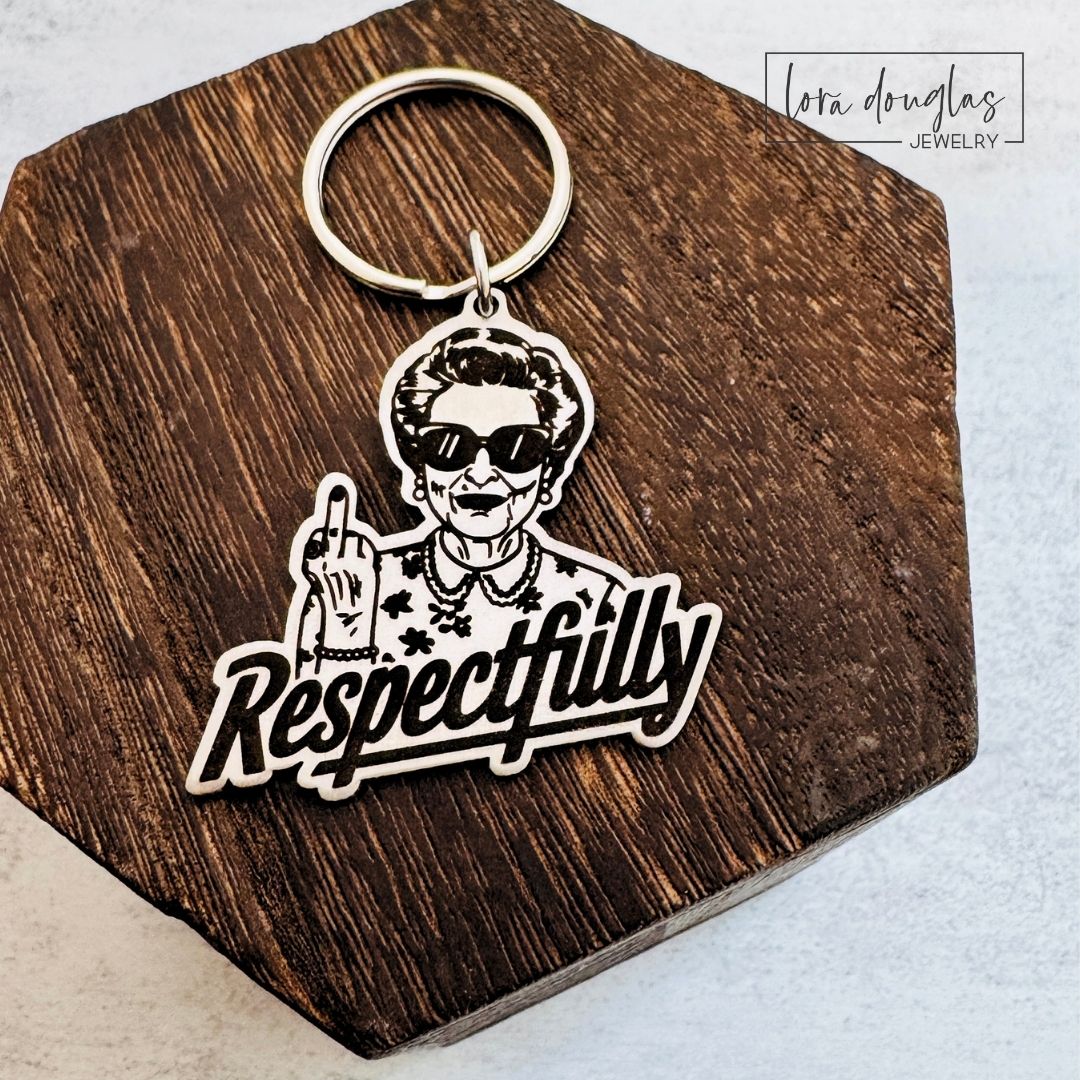 Respectfully middle finger keychain made from brushed stainless steel with a permanently laser engraved woman wearing sunglasses, shown in hand to demonstrate the approximately 1.5 inch size and included key ring.