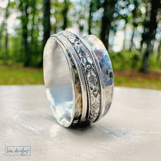 Sterling silver spinner ring with engraved floral band displayed on linen ring holder, outdoor natural light with soft forest background
