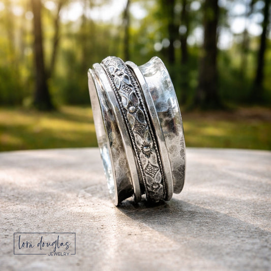 Sterling silver spinner ring with floral engraved band and wide hammered band, photographed outdoors on stone with blurred forest background