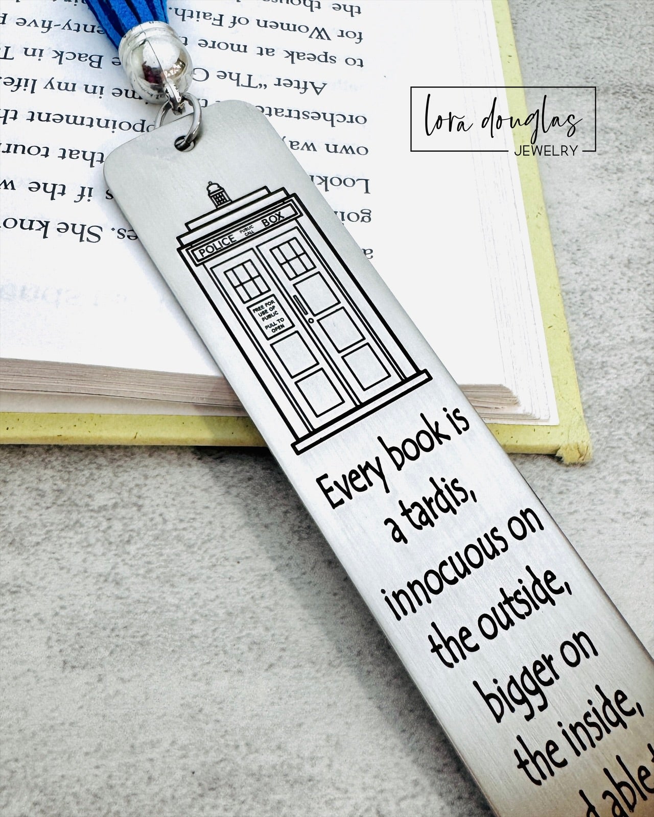 Bookmark with TARDIS design and quote on a book, featuring Lora Douglas Jewelry branding.
