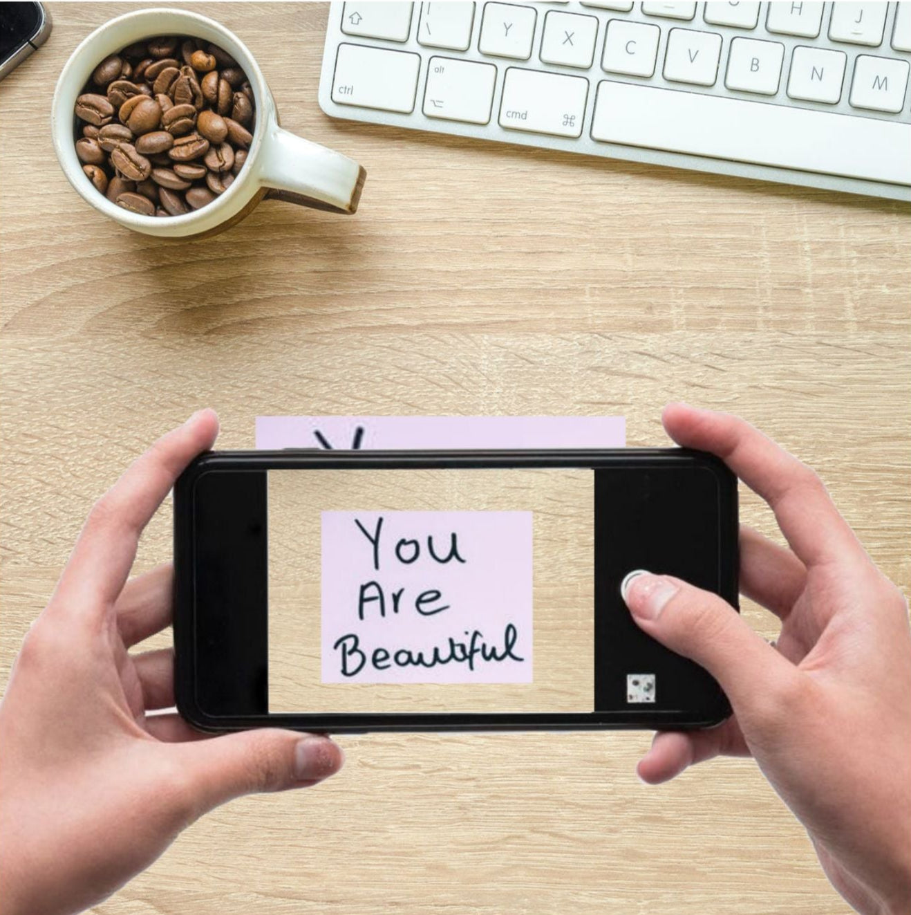 Person holding a smartphone with a 'You Are Beautiful' message on a desk with a cup of coffee and keyboard.