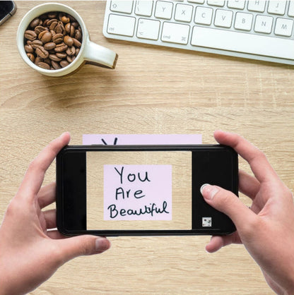 Person holding a smartphone with a 'You Are Beautiful' message on a desk with a cup of coffee and keyboard.