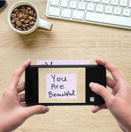 Person holding a smartphone with a 'You Are Beautiful' message on a desk with a cup of coffee and keyboard.