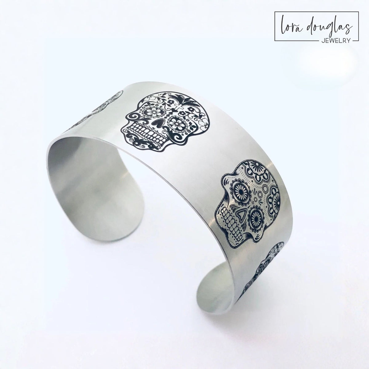 Wide sugar skull cuff bracelet in stainless steel featuring five unique engraved skull designs around the band, available in silver or gold with a matte finish for fans of bold skull jewelry and alternative style.
