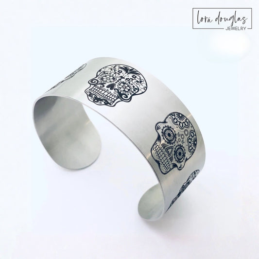 Wide sugar skull cuff bracelet in stainless steel featuring five unique engraved skull designs around the band, available in silver or gold with a matte finish for fans of bold skull jewelry and alternative style.
