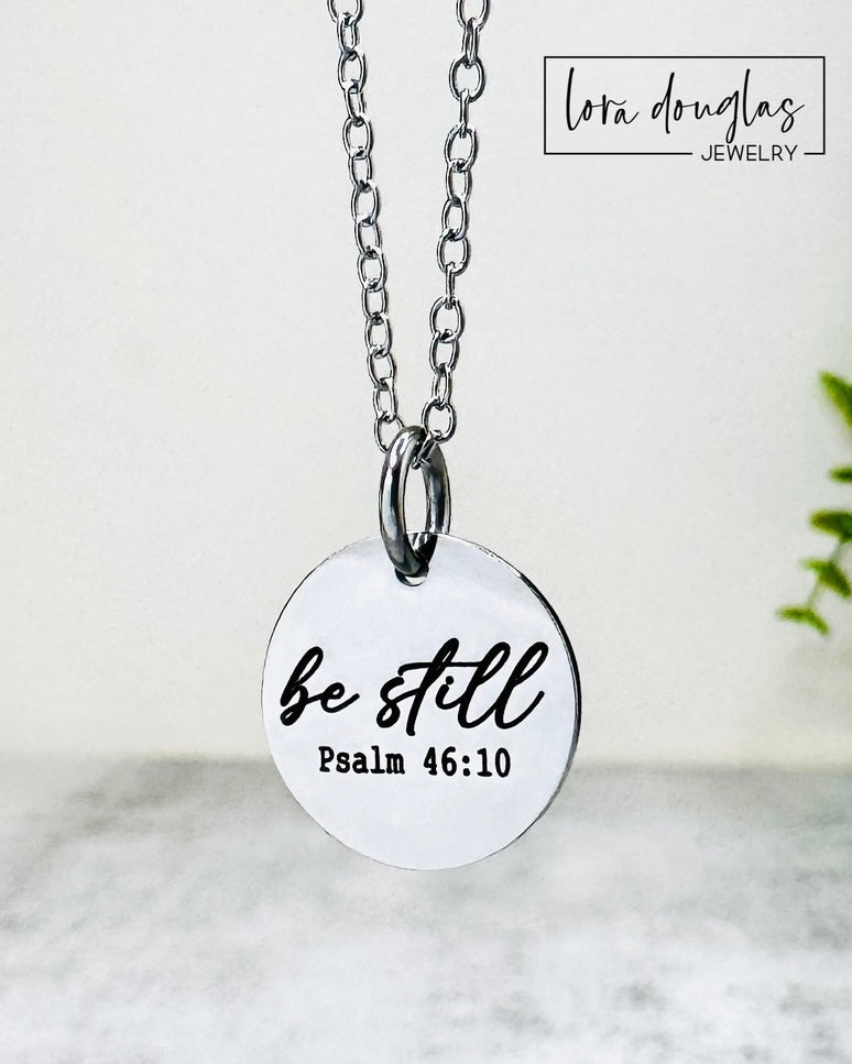 Necklace with a pendant that reads 'be still Psalm 46:10' on a light background.