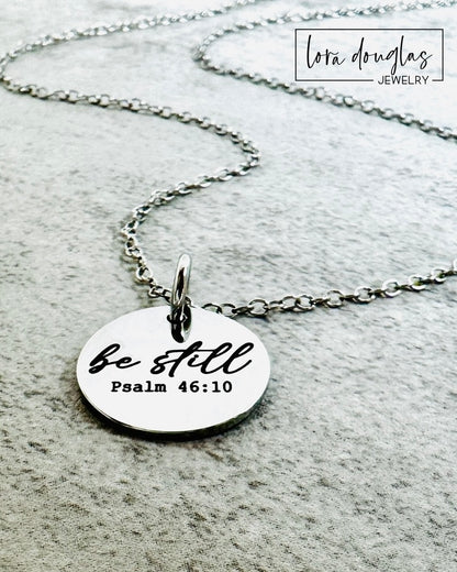 Silver necklace with a round pendant featuring 'be still Psalm 46:10' on a gray surface.