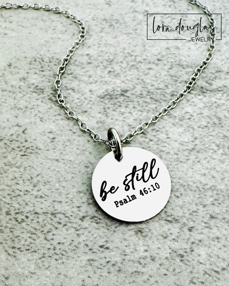 Necklace with a round pendant featuring 'be still' and 'Psalm 46:10' on a textured gray background.
