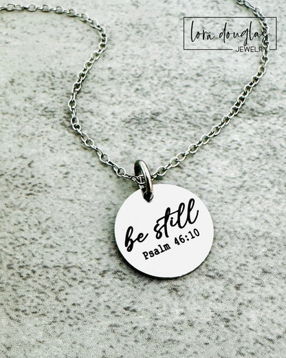 Necklace with a round pendant featuring 'be still' and 'Psalm 46:10' on a textured gray background.