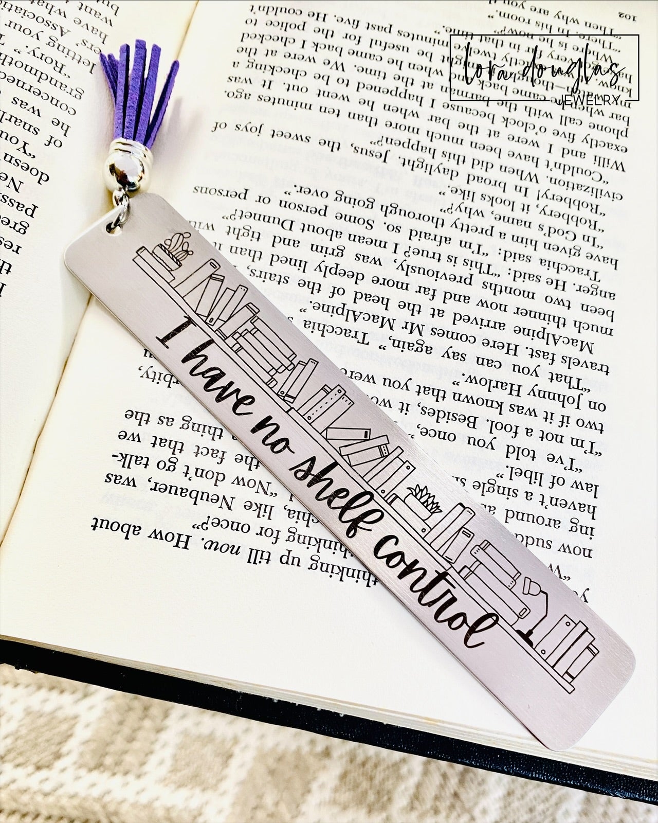 A stainless steel bookmark with an engraving that reads 'I HAVE NO SHELF CONTROL' on a bookshelf background, placed on an open book.