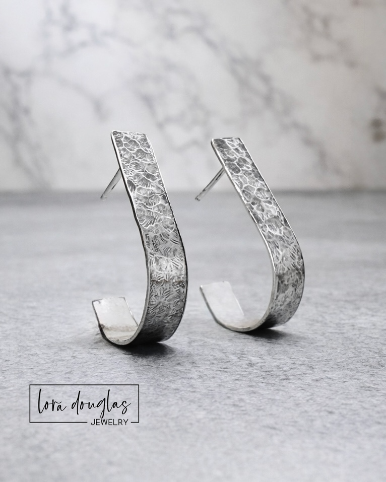 sterling Silver earrings with textured design on a gray surface, Lora Douglas Jewelry brand visible.
