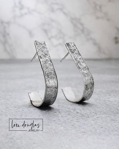 sterling Silver earrings with textured design on a gray surface, Lora Douglas Jewelry brand visible.