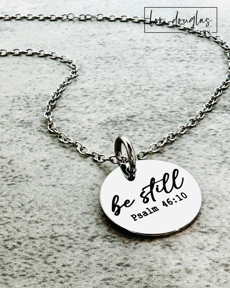 Necklace with a round pendant that says 'be still' and 'Psalm 46:10' on a textured surface.