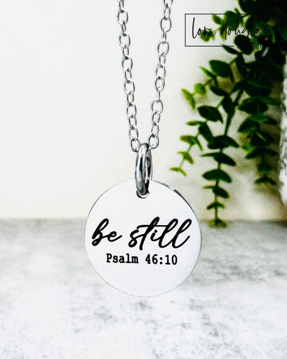 Necklace with a round pendant featuring 'be still' and Psalm 46:10 text, on a marble surface with a plant in the background.
