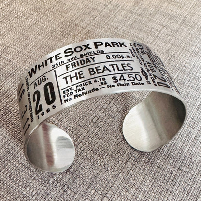 A stainless steel bracelet with engraved text representing a vintage Beatles concert ticket, stating 'White Sox Park' and the date 'Aug 20 1965' along with the band's logo and price information.