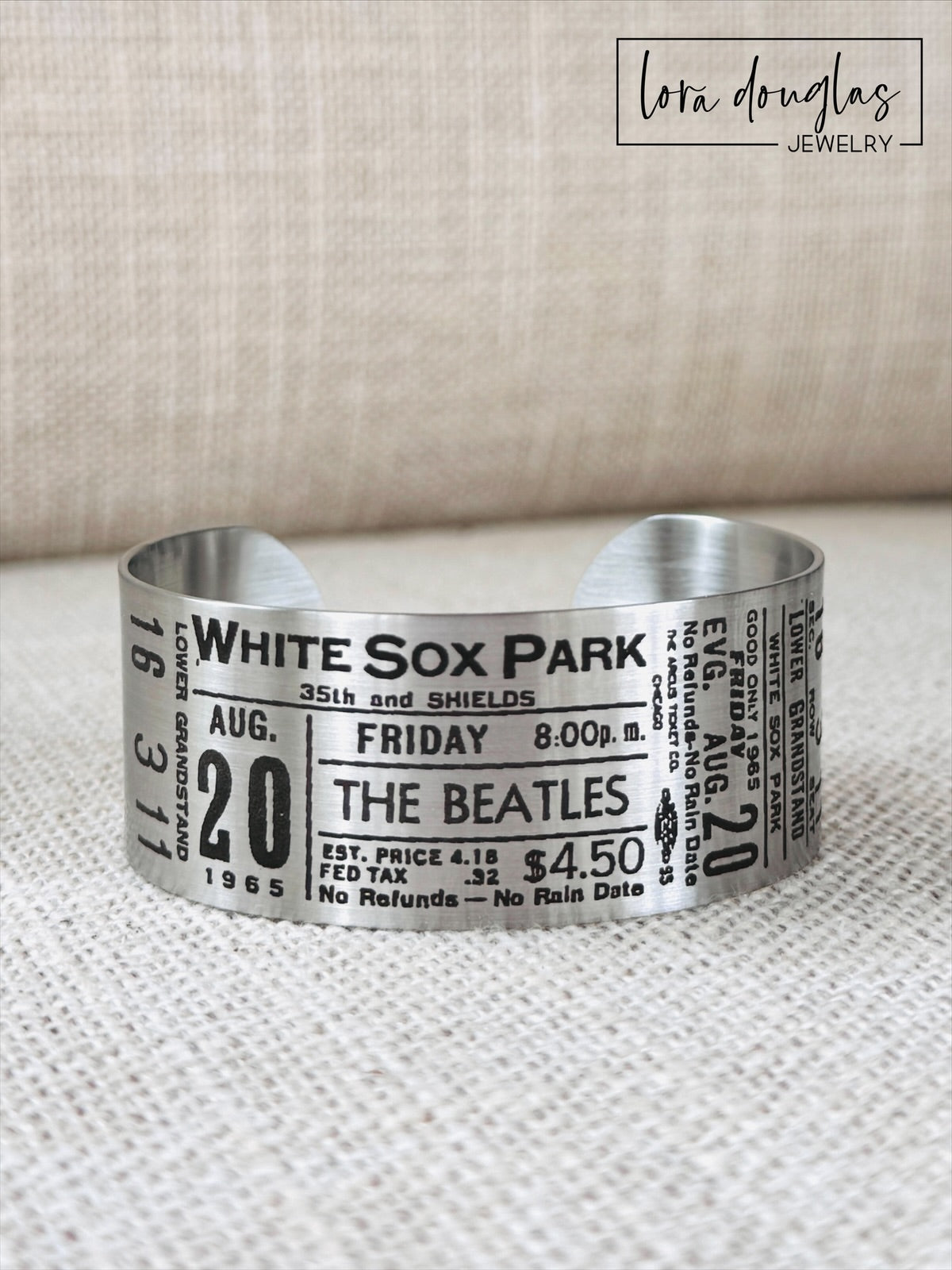 A stainless steel bracelet with engraved text representing a vintage Beatles concert ticket, stating 'White Sox Park' and the date 'Aug 20 1965' along with the band's logo and price information.