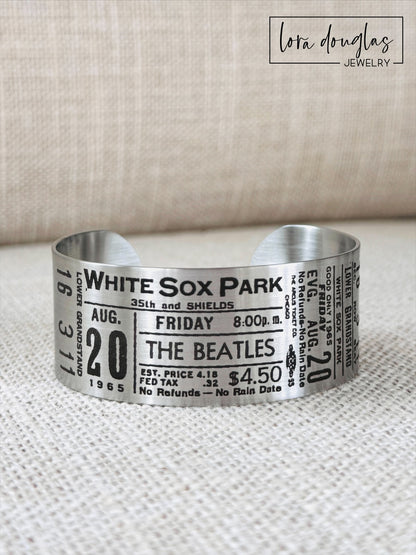 A stainless steel bracelet with engraved text representing a vintage Beatles concert ticket, stating 'White Sox Park' and the date 'Aug 20 1965' along with the band's logo and price information.