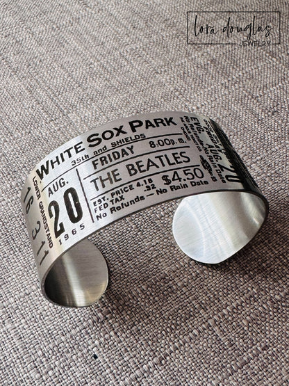 A stainless steel bracelet with engraved text representing a vintage Beatles concert ticket, stating 'White Sox Park' and the date 'Aug 20 1965' along with the band's logo and price information.