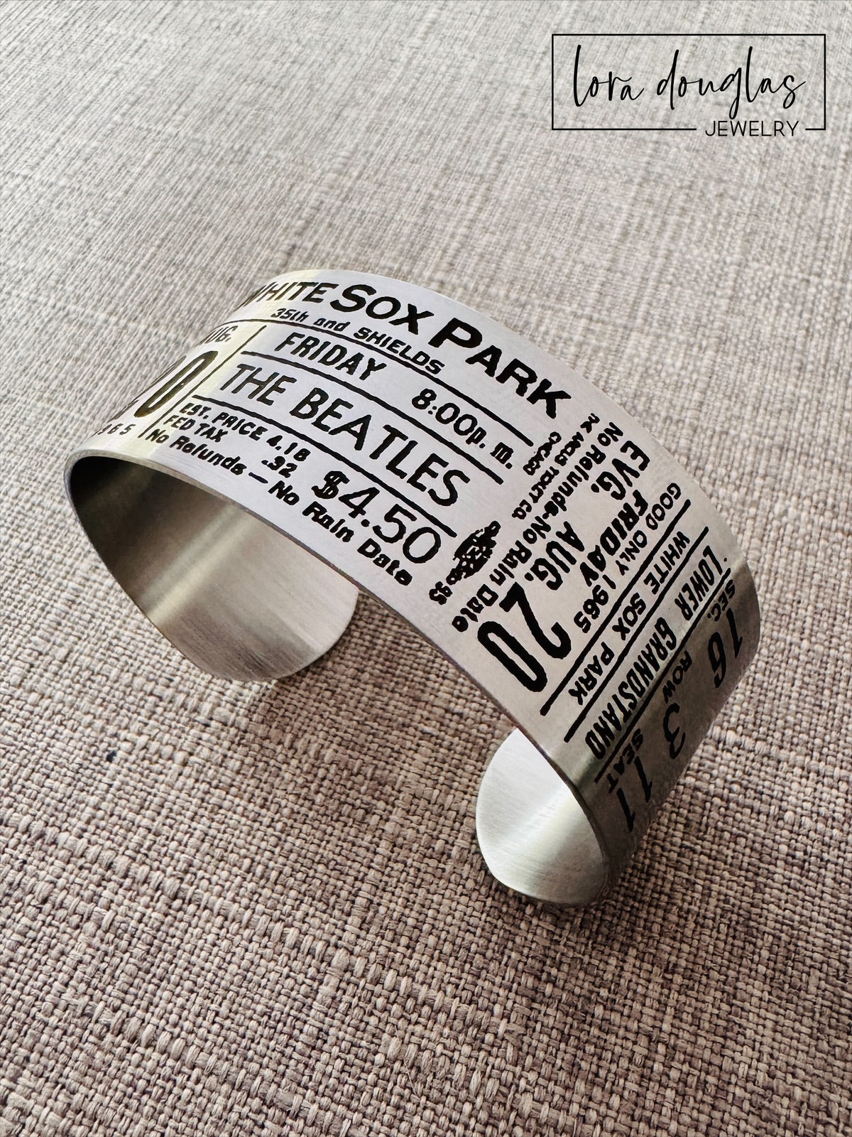 A stainless steel bracelet with engraved text representing a vintage Beatles concert ticket, stating 'White Sox Park' and the date 'Aug 20 1965' along with the band's logo and price information.