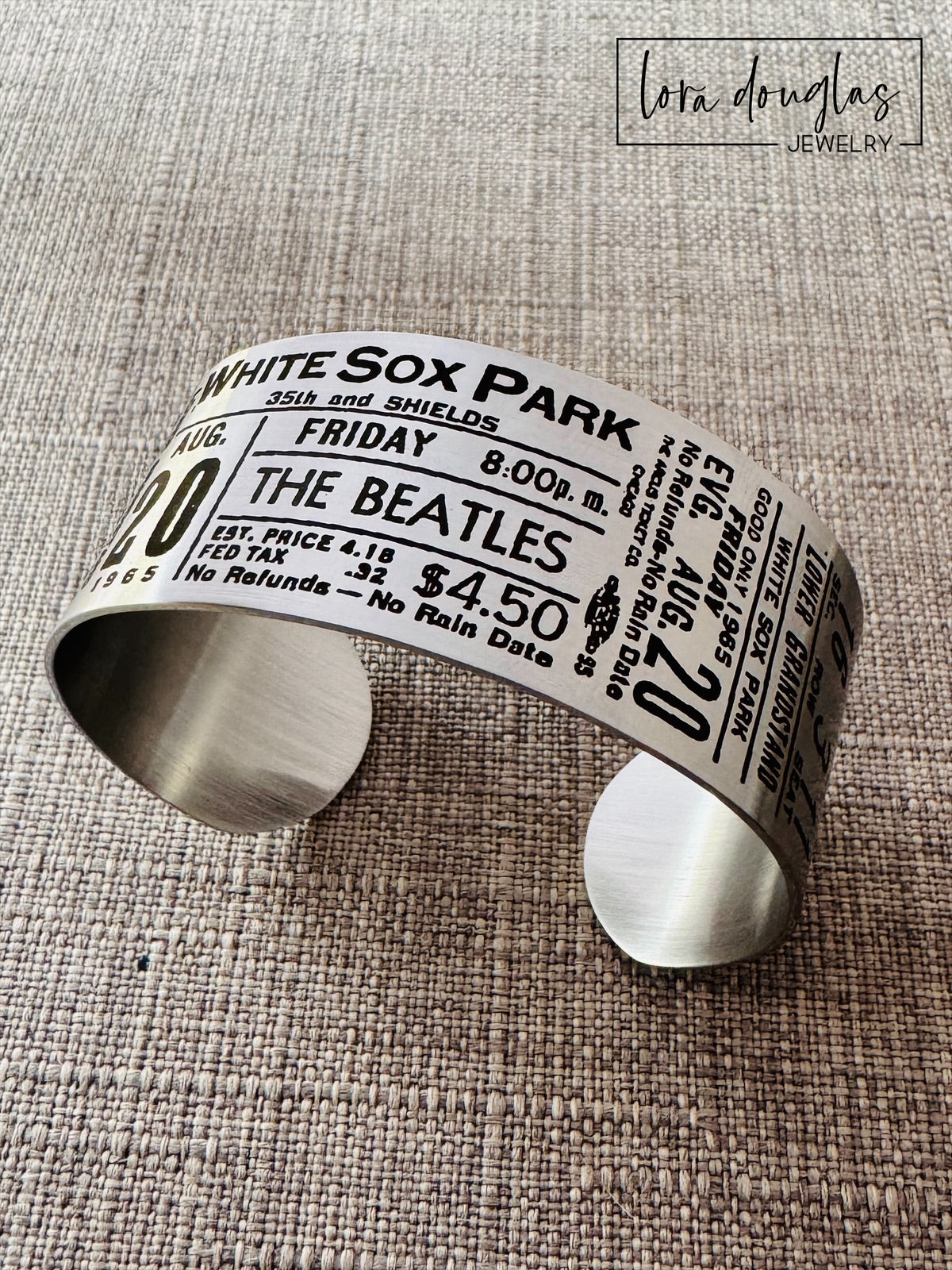 A stainless steel bracelet with engraved text representing a vintage Beatles concert ticket, stating 'White Sox Park' and the date 'Aug 20 1965' along with the band's logo and price information.