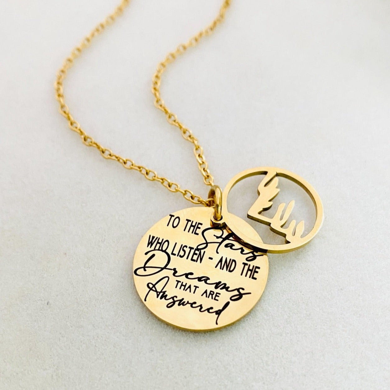 A gold stainless steel necklace with a round pendant engraved with the quote 'To the Stars who listen - and the Dreams that are Answered' and a laser cut mountain pendant.