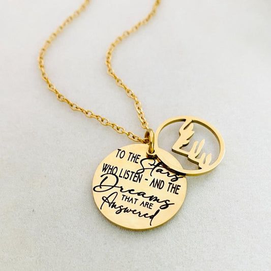 A gold stainless steel necklace with a round pendant engraved with the quote 'To the Stars who listen - and the Dreams that are Answered' and a laser cut mountain pendant.