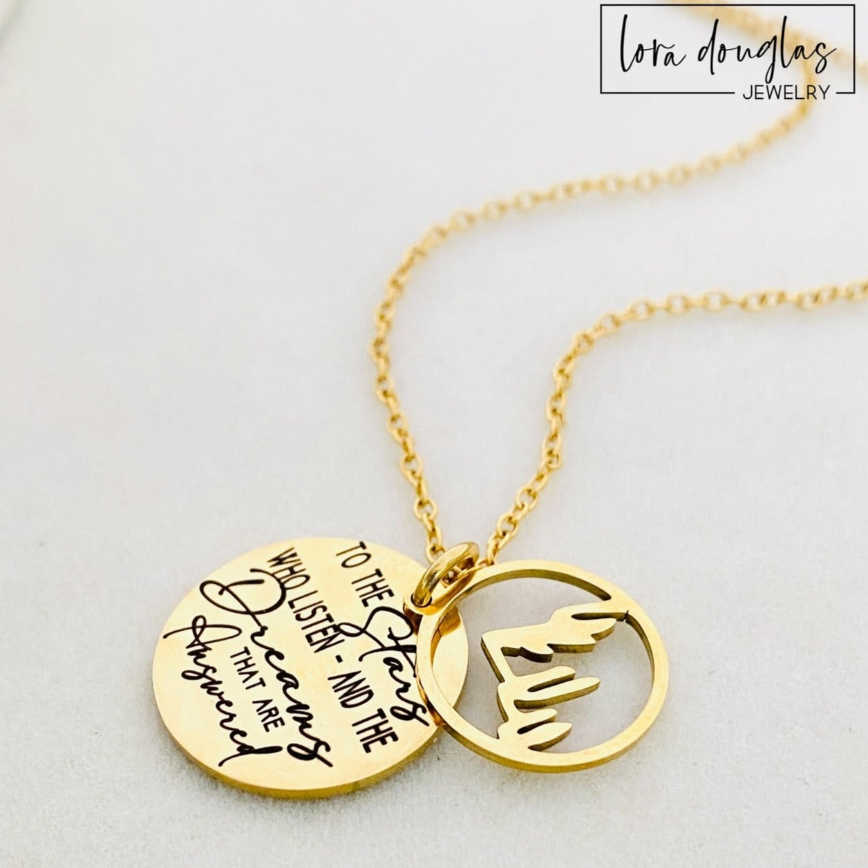 A gold stainless steel necklace with a round pendant engraved with the quote 'To the Stars who listen - and the Dreams that are Answered' and a laser cut mountain pendant.