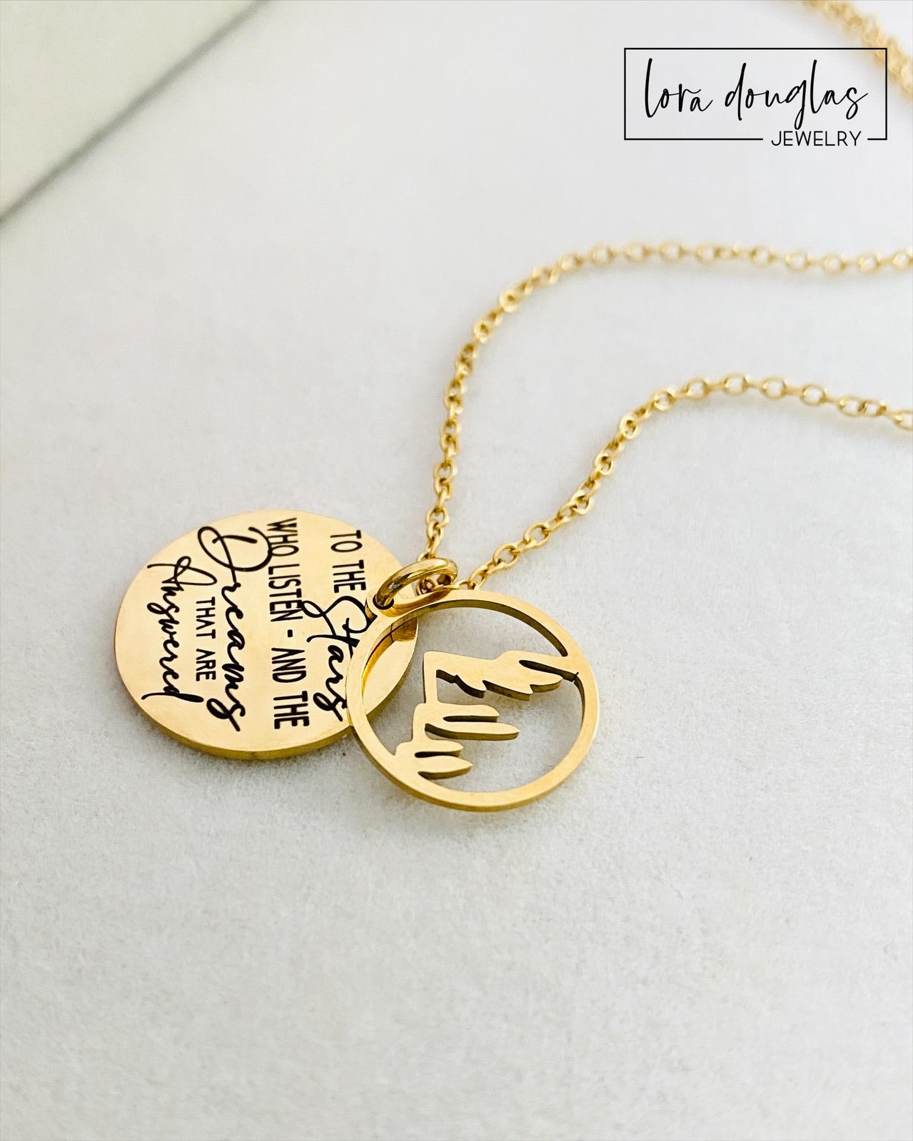 A gold stainless steel necklace with a round pendant engraved with the quote 'To the Stars who listen - and the Dreams that are Answered' and a laser cut mountain pendant.