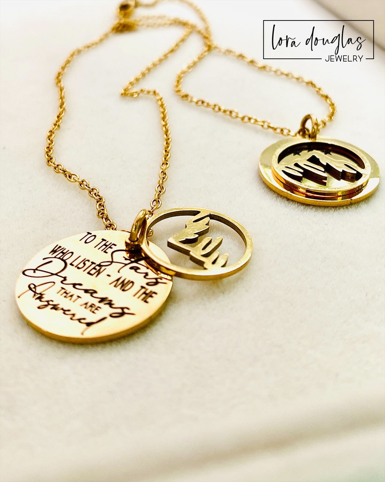 A gold stainless steel necklace with a round pendant engraved with the quote 'To the Stars who listen - and the Dreams that are Answered' and a laser cut mountain pendant.