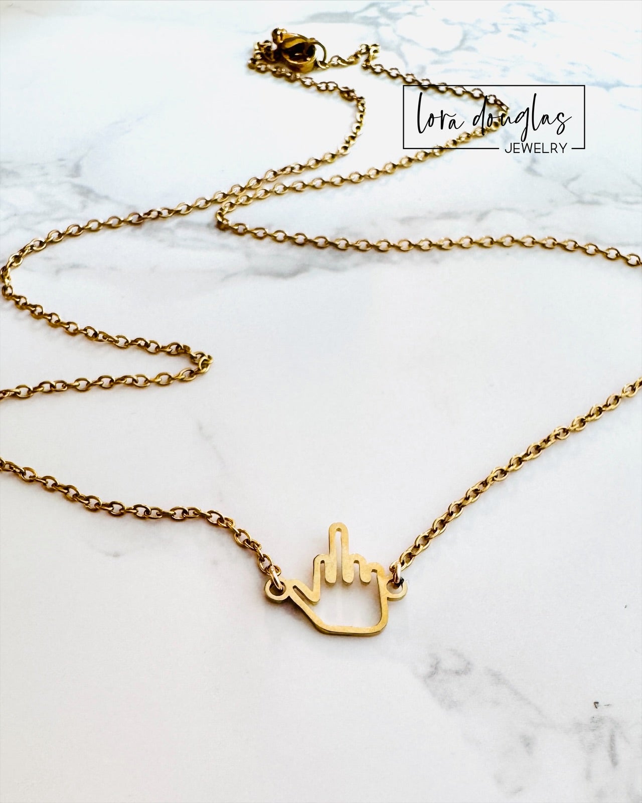 The Middle Finger Necklace by Lora Douglas features a playful middle finger charm pendant, shown on a white marble surface with the brand name displayed in the top right corner.